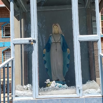 A standing statue of the Virgin Mary is enclosed within a light blue, glass-paned cabinet. The statue wears a white gown and a blue mantle, with a rosary and crucifix around her neck; her arms are extended slightly downward and outward, palms facing forward. The cabinet's left door is secured by a brass padlock. Inside the cabinet, at the base of the statue, are white and light blue artificial flowers, silver tinsel, and white sheer fabric decorations. The cabinet rests on a light blue tiled base. To the left of the cabinet, a multi-story building constructed of light brown bricks with light blue window frames and arched, grilled windows is visible. A light-colored railing is present in the far-left foreground. This scene is located in Machakos Central ward, Kenya.