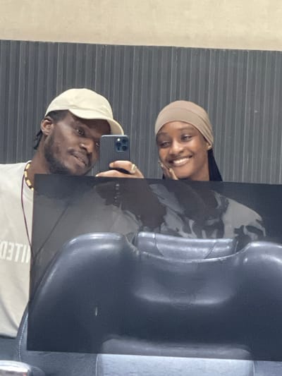 In a well-lit indoor setting, possibly a salon or studio in Guzape, Nigeria, a young African couple takes a mirror selfie. The man, wearing a light-colored baseball cap and a t-shirt with a printed design, holds a smartphone to capture the reflection. To his right, a woman in a tan headscarf smiles warmly, resting her chin on her hand. They are positioned in front of a dark, ribbed wall. In the foreground, the reflection shows the backs of two salon chairs, one dark and smooth, the other a lighter, possibly metallic shade. The overall impression is one of casual intimacy and personal documentation.