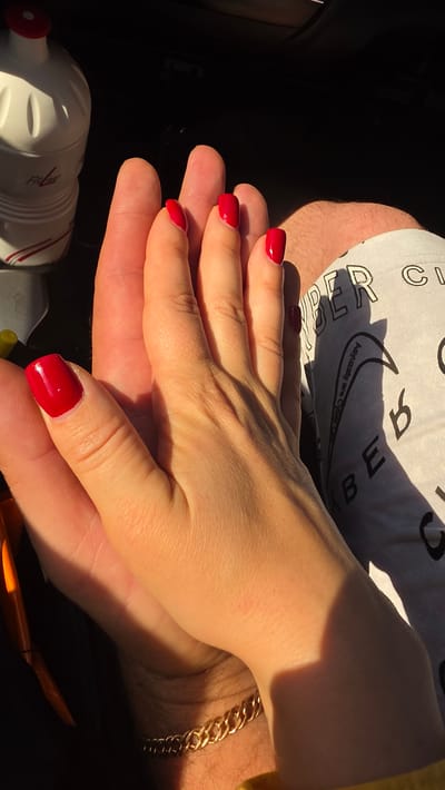 In Kruglikovo, Russia, two hands, one appearing larger and presumably male, the other smaller and female, are clasped together. The female hand, with bright red painted fingernails, rests on top of the male hand. The larger hand exhibits visible fingerprints and is adorned with a gold chain bracelet. The background is dark, suggesting an indoor setting, possibly a car interior, with a white bottle with red accents partially visible on the left. Sunlight casts a warm glow and shadows across the clasped hands, highlighting the texture of the skin and the metallic sheen of the bracelet. The female's arm is bent at the wrist, and her fingers are intertwined with the male's.