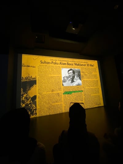 The image shows a projected image of a newspaper article in a darkened room. The article is written in Indonesian. It is titled "Sultan-Paku Alam Baca 'Maklumat 20 Mei'." The article is about a historical event in Yogyakarta, Indonesia on May 20, 1949.  There is a photograph of a man in a suit. The man is speaking into a microphone and is facing the right. The man appears to be giving a speech.  There are two silhouettes of people seated in the room, one on the right and one on the left of the projection.  The projection appears to be a classroom or other educational setting. The coordinates -7.8, 110.407 correspond to the city of Banguntapan, Indonesia.