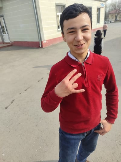 A young male, approximately a teenager, is depicted standing outdoors in Rishtan, Uzbekistan. He faces forward, looking directly at the camera with a smile. He has short, dark hair and is wearing a red long-sleeved polo shirt with a quarter-zip collar and a small dark emblem on the left chest, over a white collared shirt. He is also wearing blue jeans. His left hand is raised to chest level, forming a gesture with the index and middle fingers extended upwards, while the thumb, ring, and pinky fingers are bent. His right hand is partially obscured, appearing to be in his front right pocket.

In the background, two individuals are walking away from the camera. The closer individual is a child or young person, seen from the back, wearing a dark vest over a light long-sleeved top and dark lower garments. Behind them, another individual, also seen from the back, wears a light-colored top, possibly a jacket.

To the left and behind the main subject is a light-colored building, possibly beige, with a distinct red trim along its base. It features at least two rectangular windows with white frames and a white door on the far left. A grey downspout is visible on the building's corner. The ground is paved with light grey concrete or asphalt, showing scattered dark specks and faint shadows. The sky in the distance appears light and somewhat hazy.