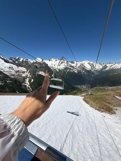 A person's hand is in the foreground, with their index finger pointing towards a ski lift in the distance. The ski lift consists of chairs suspended from cables, traversing a snowy mountain landscape under a clear blue sky. The mountains are snow-capped and rugged, with green slopes visible in the lower sections. The ski lift appears to be in operation, with several chairs visible. The hand is lightly tanned and has manicured nails. The person is wearing a white garment with ruffled sleeves. The scene suggests a sunny day, likely during the warmer months when snow persists at higher altitudes. The image evokes a sense of adventure and exploration in a mountainous environment.