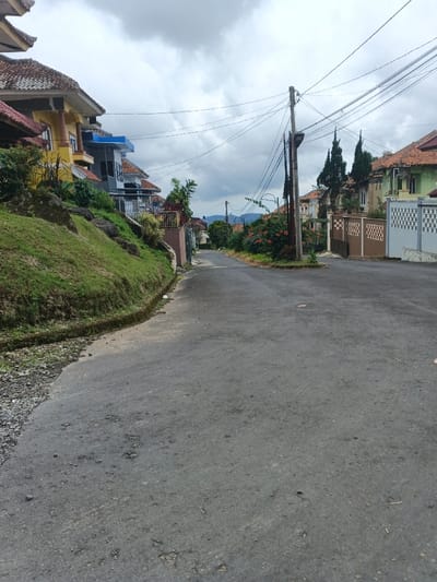 The image depicts a residential street in Pô Kô, Vietnam, under an overcast sky. The asphalt road surface is visible, showing signs of wear and minor debris, and extends into the distance, appearing to curve slightly. No individuals, vehicles, or observable actions are present.

On the left side of the street, an ascending grassy embankment supports multiple multi-story residential buildings. These houses feature varying architectural styles, with prominent tiled roofs in shades of brown and orange, and facades in colors such as yellow, white, and blue. Retaining walls and fences secure the properties along the embankment. A narrow, partially overgrown concrete drainage channel runs along the left edge of the road.

On the right side of the street, a dark utility pole stands prominently, supporting a dense network of overhead electrical and communication lines that cross the street. Adjacent to the utility pole and further down the street are additional residential properties, some featuring decorative perimeter fences and gates, including patterns such as brown checkerboard and light blue geometric designs. Greenery, consisting of bushes, flowering plants, and several tall, conical evergreen trees, is interspersed between the properties. Distant hills are visible in the background beneath the cloudy horizon.