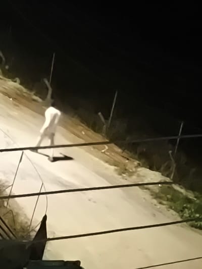 The image depicts an outdoor scene at night in Nicosia, Northern Cyprus / Cyprus. A single individual, an adult male, is visible on a light-colored, unpaved or gravel road. He is wearing light-colored clothing, likely a white shirt and trousers, and is captured in mid-motion with his arms raised and bent. An elongated, distinct shadow from the individual is cast to his left on the road, suggesting an artificial light source from the right-front relative to the figure.
The environment includes several dark, vertical utility poles with overhead lines, positioned intermittently along the sides of the road. The ground bordering the road shows darker patches of earth and sparse vegetation. Multiple dark, horizontal lines, consistent with electrical or communication wires, traverse the foreground, partially obscuring the lower section of the frame. The background is uniformly dark, indicating a lack of significant ambient light. A dark, indistinct object or structure is partially visible in the bottom-left corner of the image. The overall image quality is characterized by blur and grain, typical of low-light photography.