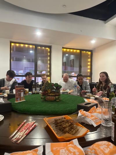 The scene depicts six individuals, five young adults and one middle-aged male, seated around a large, dark, circular dining table in a brightly lit indoor restaurant in Huangdao District, China. Overhead lighting and decorative string lights outlining two large windows illuminate the space, revealing blurred external reflections.
A central green circular mat on the table supports a multi-tiered decorative structure resembling an ornate miniature building or landscape feature. Food items include numerous skewers of grilled meat on an orange tray, various dishes on individual plates, and packaged chopsticks. Beverages consist of beer bottles, including one identified as Tsingtao Pure Draft, and water bottles, alongside empty and full clear drinking glasses. A black tissue box and a stand-up promotional card with Chinese characters, including "扫码支付" (scan code to pay), are also on the table. Small crumpled orange-and-white paper wrappers are scattered on the table surface.
The individuals are casually dressed. The person on the far right, wearing a plaid shirt, is actively eating with chopsticks. The middle-aged male in a white shirt is looking down at his plate. Other individuals are generally observing the table or their food, consistent with a group meal. No direct interpersonal interactions are distinctly captured.