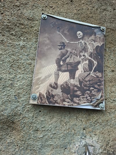A sepia-toned print, depicting two skeletons and a human in mountaineering attire, is affixed to a textured concrete wall. The print shows one skeleton holding a camera aloft, as if taking a selfie, while another skeleton stands beside it. A man in a hat and mountaineering gear, carrying a large bag, is positioned in the foreground. The print is secured to the wall by four metal screws, one of which is visible in the bottom right corner, partially obscuring the text "MEMENTO MORI" at the bottom of the print. The wall appears weathered and the print itself shows signs of damage and peeling. The overall scene suggests an outdoor setting, possibly a tourist trail or viewpoint, in Děčín, Czechia.