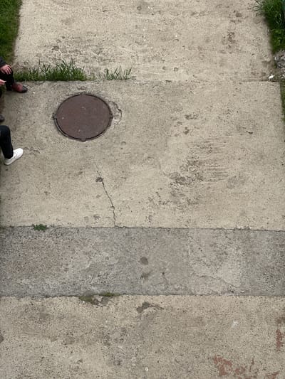 The image presents an overhead view of a weathered concrete surface, divided into distinct sections. A circular manhole cover is visible in the lower left quadrant. Vegetation, including grass, grows along the edges and between the concrete sections. Two individuals are partially visible at the bottom of the frame, one wearing a dark jacket and brown shoes, the other wearing black pants and a white shoe. The environment suggests an urban or semi-urban setting, possibly in Sayansk, Russia.