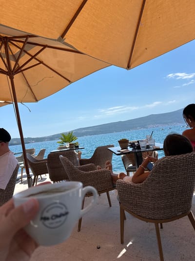 This image depicts a person's hand holding a white espresso cup with a logo that appears to say "Mr. Cha Cha" in a circular design. The cup is filled with a dark liquid, likely espresso. In the background, an outdoor seating area is visible with wicker chairs and tables. Beyond the seating area, a vibrant blue sea stretches out, meeting a distant, tree-covered coastline under a clear blue sky. The overall setting suggests a cafe or restaurant situated near the water, possibly in a Mediterranean or coastal region. The specific location is identified as Donja Lastva, Montenegro, a coastal settlement.