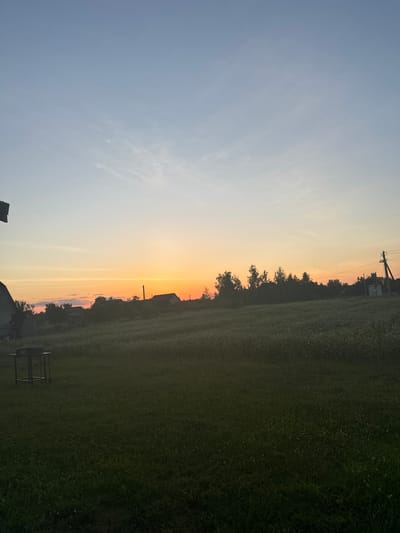 This media file captures a serene, rural landscape at dusk in Chemerne, Ukraine. The image is oriented vertically, with the sky taking up the left half and the land the right.
The sky transitions from a pale blue at the top to warm hues of orange and yellow near the horizon, indicating the time of day is sunset. Wispy clouds are scattered across the upper part of the sky.
The right side of the image shows a vast, green field, possibly farmland, stretching into the distance. A line of trees borders the field along the horizon, with silhouetted houses and structures interspersed among them. A prominent utility pole with electrical wires stands near the center of the image, a man-made element in the natural setting. In the foreground on the right, a dark, metallic object resembling a grill or a metal frame is visible. In the lower left corner, the roof of a building or structure is partially visible.
The overall mood is peaceful and tranquil, with the soft glow of sunset creating a warm and inviting atmosphere. There is no visible text in the image.