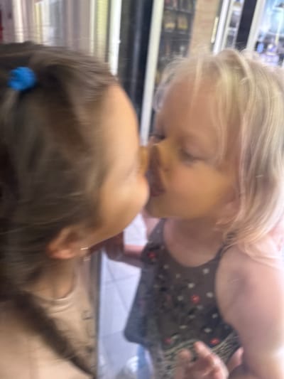 The image shows a close-up, blurred view of two young children interacting. The child on the left has dark hair pulled back with a blue hair tie. The child on the right has blonde hair and is wearing a dark sleeveless dress with a floral pattern. The children appear to be very close, possibly sharing a kiss or hug, with their faces touching. The background is out of focus but suggests an indoor environment with vertical elements that could be shelving or part of a structure. The overall impression is a moment of affection captured in a casual setting. The location is stated as Yalta, Ukraine.