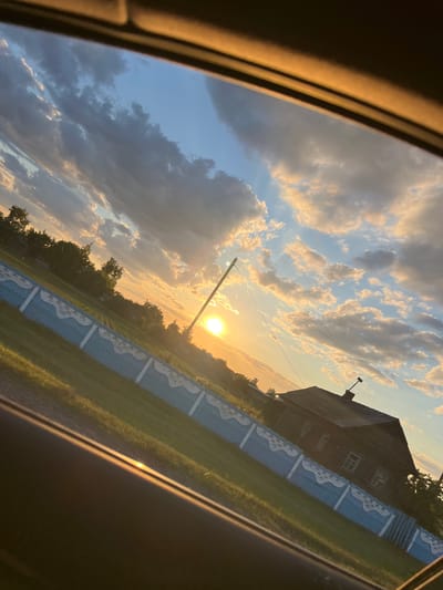 The image captures a rural scene at sunset, viewed from inside a vehicle. The sun, a bright orb, is low in the sky, casting a warm golden glow across the landscape. Wispy clouds are scattered across the blue sky, tinged with the colors of sunset.

In the foreground, a dark car interior frames the view, with a curved, illuminated edge hinting at the window or dashboard. A light blue and white picket fence runs diagonally across the middle ground, separating a grassy area from a darker area beyond. Behind the fence, a traditional wooden house with visible windows and a chimney stands prominently. Trees, some lush and green, others appearing more sparse, dot the landscape around the house. Power lines are also visible stretching across the sky.

The overall mood is peaceful and serene, evoking the quiet beauty of a countryside evening. The time of day is clearly sunset, indicated by the low sun and warm lighting. While no specific event is depicted, the scene suggests a moment of observation and perhaps travel, as the viewpoint is from within a vehicle. The location context of Азарыцкі сельскі Савет, Belarus, suggests a setting in rural Belarus. There is no visible text in the image.