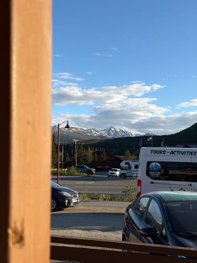 The image shows a scene with mountains in the background, a blue sky with clouds, and several vehicles parked on a paved surface. A large white tour bus with the text "TOURS - ACTIVITIES" and a logo that says "DENALI PARK ADVENTURES" is visible. Next to it, a dark-colored car is parked, and further down the road, a white camper van and another vehicle are present. In the foreground, a wooden structure partially obstructs the view. A red lamp post with a distinctive shade stands in front of the mountains. The snow-capped mountains suggest a mountainous terrain, possibly in a park. The time of day appears to be daytime, with bright sunlight casting shadows. The weather seems clear with some clouds. The text "DENALI PARK ADVENTURES" indicates the location is likely Denali National Park in Alaska, and the presence of tour buses suggests tourism activities. There are no people clearly visible in the image.