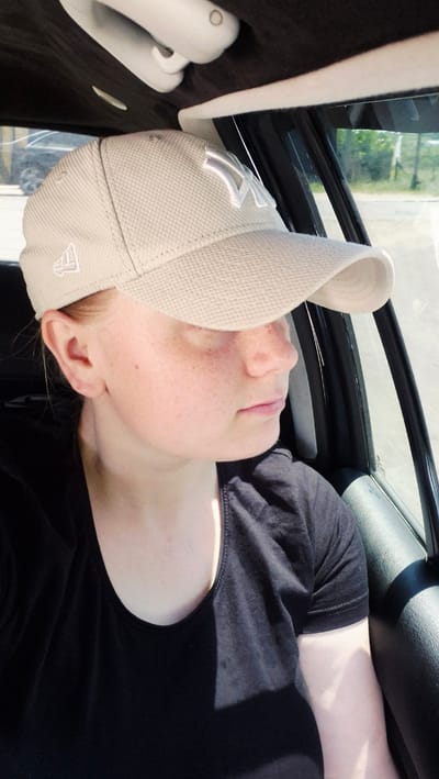The image shows a young woman sitting in the passenger seat of a vehicle. She is wearing a tan baseball cap with a white logo and a black t-shirt. She has fair skin with freckles and her hair is blonde. She is looking out the window of the car. The interior of the car is dark, with a beige headliner and black trim visible. The exterior of the car shows greenery and a road, suggesting an outdoor setting. The image is taken from a slightly elevated angle, looking down at the woman. The lighting suggests it is daytime. The image does not provide any information about the city of Piatra Albă, Moldova, or any specific location within it.