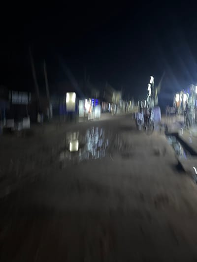 The image shows a blurred, sideways view of a street in Eziobodo, Nigeria, at night. Illumination comes from streetlights and building lights, creating streaks due to motion blur. A dark, unlit area occupies the left side of the image. On the right is a dark colored wall. Other details are unidentifiable due to the blur. No people are clearly visible.