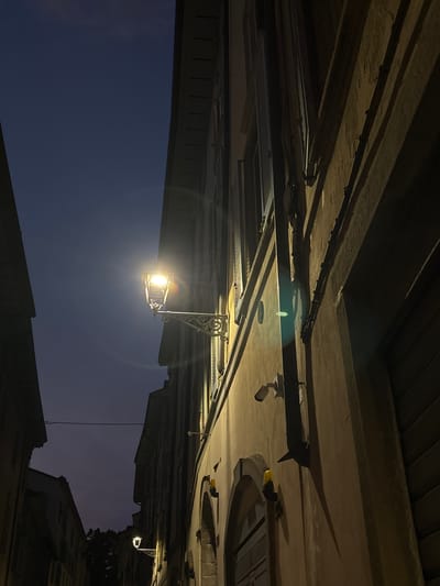 A street lamp illuminates a narrow, darkened street in Bergamo, Italy, at twilight. The lamp, with an ornate metal fixture, casts a warm glow on the building facade. The building itself is old, with visible architectural details and shutters on the windows. Shadows are cast by the building and other unseen objects, creating a dramatic contrast between light and dark. The sky is a deep, dusky blue, indicating the transition from day to night. There are no people visible in the frame.