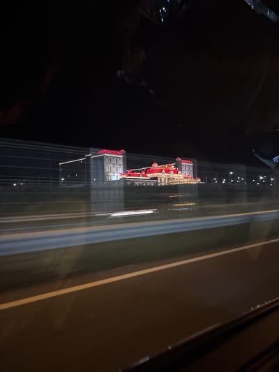 The image captures a large, illuminated building complex at night, viewed from the interior of a moving vehicle on a multi-lane road in Kapitan Andreevo, Bulgaria. The primary subject is a multi-sectioned structure featuring distinctive red, ornate roofs and light-colored facades, brightly lit by artificial white and yellow light, highlighting its architectural details. The building appears to be part of a larger complex, potentially administrative or commercial, common at a major border crossing.
In the foreground, a dark asphalt road surface is visible, marked by a single yellow lane line. A blurred grey barrier or divider runs horizontally across the mid-ground, indicative of a highway or major thoroughfare. Horizontal streaks of light and blur in the middle distance reinforce the impression of motion. The upper portion of the frame shows dark reflections from inside the vehicle, possibly including elements of the dashboard or interior ceiling. The background is largely dark, consistent with nighttime, with scattered distant lights from additional streetlights or structures visible.
