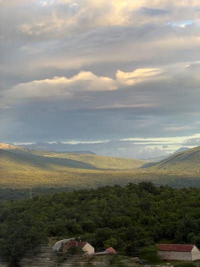 This landscape photograph presents a vast natural scene of rolling hills and mountains covered in dense forest, under a dramatic, partially clouded sky. The image appears to be taken during late afternoon or early evening, as indicated by the warm, golden light illuminating parts of the clouds and distant hills, suggesting the sun is low on the horizon.

The upper portion of the frame features a dynamic sky with large, textured clouds. Some of these clouds catch the golden sunlight, appearing bright and ethereal, while others remain in deeper grey tones, creating a striking contrast. Below the sky, successive layers of tree-covered hills and mountain ranges recede into the distance, displaying various shades of green from the extensive vegetation. The dense forestry suggests a rich, untamed natural environment.

On the right side of the image, two structures are visible. A larger, light-colored building with a reddish-brown tiled roof stands in the mid-ground, resembling a traditional rural dwelling or barn. Closer to the foreground and slightly to its left, a smaller building with a similar roof can be seen, partially nestled among the trees. A narrow, dark road or path winds past this smaller building and extends into the forested area. A utility pole with visible lines is also present in the middle distance, integrated within the tree line.

No people or specific activities are depicted, giving the scene a tranquil and expansive feel, emphasizing the untouched beauty of the natural world. The rugged terrain and traditional architecture align with the context of a mountainous region like Šipačno, Montenegro. There is no visible text in the image.