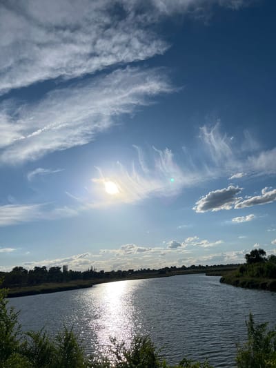The image shows a wide, calm river flowing through a landscape with trees and vegetation along its banks. The sun is shining brightly, creating a strong reflection on the water's surface. The sky is a clear blue with scattered, wispy white clouds. The scene appears to be a natural outdoor setting, possibly a park or a rural area. The crops suggest the location is Shyroka Dacha, Ukraine.