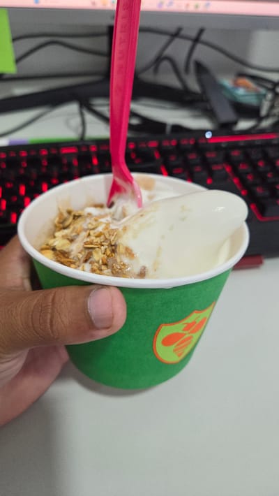A hand holds a green paper cup containing what appears to be vanilla ice cream with granola. A pink plastic spoon is partially visible inside the cup. In the background, a computer monitor and a keyboard with red backlighting are present. The location of the scene in Sanctorum, Mexico, cannot be determined from the visual information provided in the image.