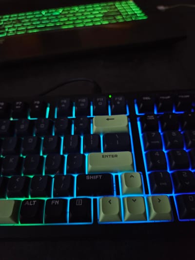 This is a close-up, low-angle, dimly lit photograph of a black mechanical keyboard with multi-colored backlighting. The top rows of keys are illuminated with green and blue light, while the main body of the keyboard, particularly the central and lower sections, is predominantly lit with bright blue light. The keys themselves are black with white lettering. Several keys are highlighted with different colored backlighting, including a bright green "Enter" key and a pale green arrow key to its left. The top portion of the image shows a blurred background of more green-lit keys, likely from another keyboard or a different section of the same keyboard. There are no people or animals visible in the image. The setting appears to be indoors, likely in a room with low ambient light, as evidenced by the dominance of the keyboard's backlighting and the dark surroundings. There is no information in the image to suggest a location in the city of Versailles, France.