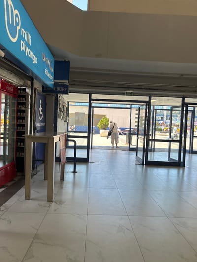 The image shows the interior of what appears to be a transportation hub or public building in Bayrampaşa, Turkey. In the foreground, a white table with four legs stands on a tiled floor with a marble pattern. To the left of the table is a red Coca-Cola refrigerator. Behind the table, there is a blue door with a white wheelchair accessibility symbol and the text "ENGELLİ TUVALETİ" (Accessible Toilet) written below it. Further back, a person in traditional white clothing is walking away from the camera, carrying a bag over their shoulder. The person is walking towards a set of open glass doors, beyond which is an outdoor area with greenery, people, and vehicles. An illuminated green sign with "EXIT" and "ÇIKIŞ" (Exit in Turkish) is visible above the glass doors. A blue sign with "MİLLİ PİYANGO" (National Lottery) and "BİZİM BÜFE" (Our Kiosk) is mounted on the ceiling. The environment appears to be brightly lit by natural sunlight.