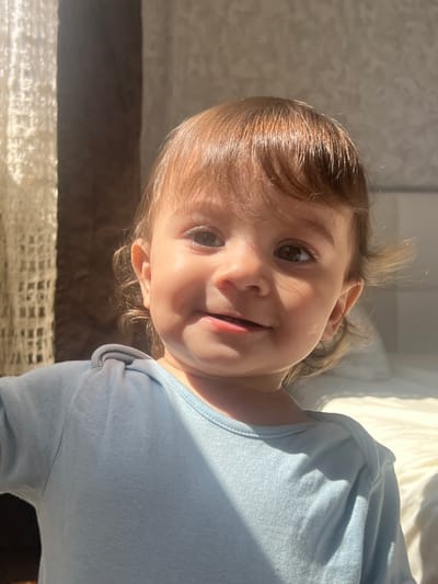 A young child with light brown hair and brown eyes is tilted towards the camera, smiling slightly. The child is wearing a light blue garment. The background is blurred and suggests a domestic interior with sunlight streaming in. There is no definitive evidence of a specific location in Elena, Bulgaria.