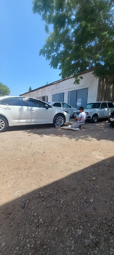 A young male kneels on a mat in front of a white sedan in an outdoor lot. He is wearing a light-colored t-shirt and gray pants. In front of him, he appears to be working on something near the front wheel of the car. The car is parked on a gravel surface in front of a long, white building with blue garage doors. The building has some writing on it in Cyrillic script. To the right of the sedan are two other white cars. A tree with green leaves provides shade over a portion of the scene. The sky is clear and blue. This scene is located in Almalyk, Uzbekistan.