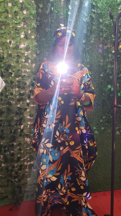 A dark-skinned woman stands in front of a backdrop of artificial green ivy and grass. She is wearing a black dress with a floral pattern of blue flowers and yellow accents. She also has a matching headscarf. The woman is holding a smartphone in both hands, and the screen is illuminated, casting a bright light on her face and the front of her dress. A vertical black pole, possibly part of a photography setup, is visible to the right of the woman. The floor is covered in red carpet. This image was taken in the city of Kinkinau, Nigeria.