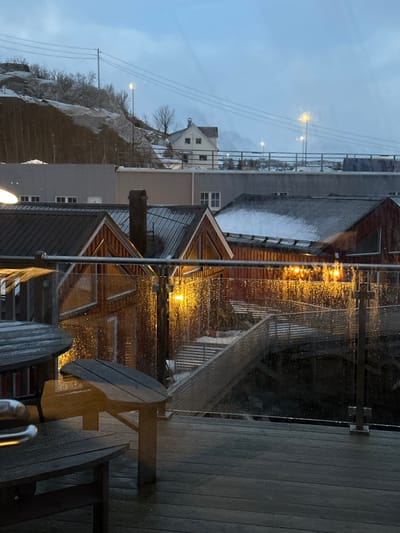 A rainy view showcases buildings with metal roofs and wooden siding. Streetlights cast a glow on the wet surfaces. In the foreground, an outdoor table and chairs sit on a wooden deck behind a glass railing. A hillside with sparse vegetation is visible in the distance under an overcast sky.