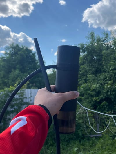 A hand in a red long-sleeved shirt with a white emblem is holding a black hookah stem. The background shows a bright blue sky with white clouds, lush green trees, and a white fence. The scene appears to be outdoors, possibly in a backyard or a park area in Vasilyevo, Russia, given the context. The lighting suggests it is daytime.