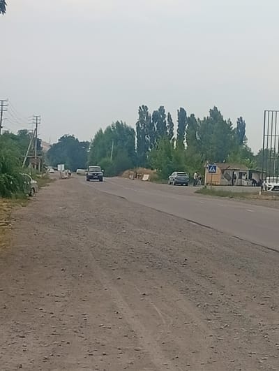 In Tash-Moinok, Kyrgyzstan, a two-lane asphalt road stretches through a rural or semi-rural landscape. The road is bordered by dirt shoulders and sparse vegetation. Several vehicles are present, including a dark SUV traveling on the road, a white car parked on the left shoulder, and a silver SUV near a small roadside structure. This structure appears to be a makeshift stall or shop with people standing near it. Power lines and poles are visible on the left side of the road, and tall trees line the right side. The sky is overcast and hazy.