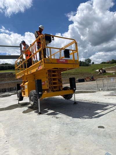Two construction workers, wearing hard hats and high-visibility vests, stand on the platform of a yellow scissor lift at a construction site. One worker is looking down at a device, possibly a tablet or control panel, while the other is looking towards the camera. The scissor lift is positioned on a concrete surface, with its stabilizers deployed. In the background, there is a hillside with trees and some buildings. The sky is blue with scattered white clouds. Several excavators and construction barriers are visible in the middle ground, suggesting ongoing construction activity. The scene appears to be a sunny day, as indicated by the shadows cast by the scissor lift and its occupants. The location is identified as Janowice, Poland.