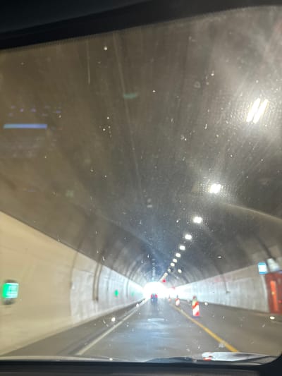 The image captures a view from inside a vehicle driving through a brightly lit tunnel. The tunnel walls are textured and light-colored, with a series of illuminated fixtures running along the ceiling. Several green rectangular signs are visible on the left wall, and at least one blue sign with white text is present on the right. Traffic cones with red and white stripes are positioned along the right side of the road, and a yellow line marks the center. A vehicle is visible in the distance ahead. The windshield of the vehicle shows numerous specks of dirt or dust. This scene is characteristic of the highway tunnel sections in the municipality of Waldhufen, Germany, specifically the A4 autobahn which passes through the area, known for its modern infrastructure including tunnels.