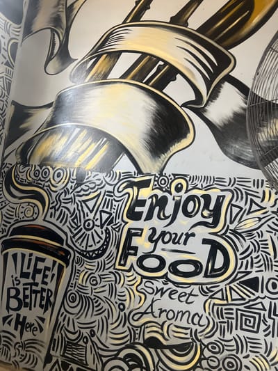 The image displays a vibrant mural, likely adorning a wall in a cafe or restaurant in Barako, Nigeria. The artwork is characterized by a black, white/light gray, and gold color scheme and combines two distinct styles.
On the left side, the mural features large, stylized illustrations of kitchen utensils, including a prominent fork and other swirling, ribbon-like forms that suggest movement. These elements are rendered with shading in black, white, and gold, giving them a metallic or lustrous appearance. A portion of what appears to be a ceiling fan is visible at the top edge of this section.
The right side of the mural is densely packed with intricate, abstract doodle-style patterns consisting of lines, swirls, geometric shapes (like triangles and squares), and circles. Interspersed within these patterns are several phrases. The most prominent text reads "Enjoy your Food," with "Enjoy" and "Food" in large, bold, black lettering, and "your" in a smaller, flowing script. Below this, in a more delicate cursive, is "Sweet Aroma." Further down and to the right, within a stylized cup or mug design, the words "LIFE IS BETTER HERE" are clearly visible, rendered in black block letters with "is Better" in a slightly smaller font.
No people or activities are depicted; the image solely captures the static artwork. The combination of food-related imagery and positive messaging, alongside the busy yet harmonious graphic design, suggests a warm and inviting atmosphere.