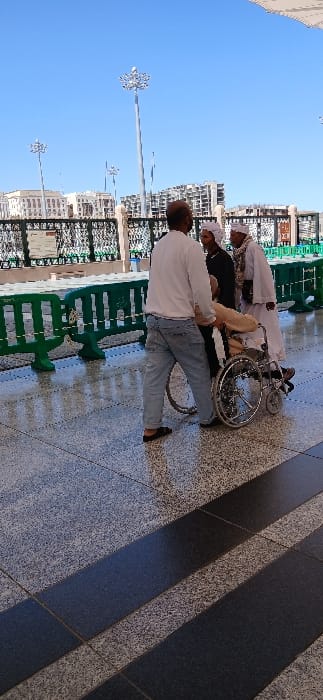The image displays an outdoor scene in Medina, Saudi Arabia, specifically in a tiled plaza or walkway near Al-Masjid an-Nabawi. Four individuals are present under a clear blue sky. In the foreground-center, an adult male, seen from the back, wears a white long-sleeved shirt, light blue jeans, and dark sandals. He is actively pushing a manual wheelchair occupied by a person dressed in light-colored traditional garments, including a head covering. To the right of the wheelchair, an adult male wearing a dark top, a white head covering, and a white shawl stands, facing towards the wheelchair. Further to the right, another adult male in a light-colored thobe and a white head covering stands, also oriented towards the central group. The ground is paved with large, polished tiles featuring speckled light and dark gray patterns, with diagonal strips of solid dark gray. A line of dark green plastic barriers extends horizontally across the mid-ground. The background reveals several tall, ornate silver streetlights and modern multi-story buildings, some with visible scaffolding, against a bright, cloudless sky. The scene is illuminated by natural daylight, casting shadows.