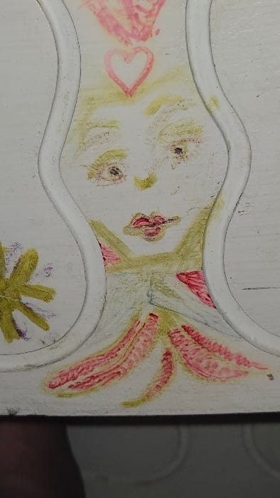 The image displays a close-up, vertical view of an abstracted, stylized humanoid figure drawn in a crayon-like medium on a textured white surface, possibly a decorative panel or piece of furniture, situated in Lompoc, United States.

The central figure features a pale yellowish-green head with two dark, roughly circular eyes exhibiting subtle red detailing, and a small, distinct red mouth. Above the head, a pink outlined heart is visible, topped by a broader, scribbled pink area. The figure's body is indicated by further yellowish-green coloration, with multiple irregular, elongated shapes in red and pink extending downwards from the mid-section, some appearing ribbed or textured.

The drawing is partially framed by undulating, raised white curvilinear borders that run vertically along both sides, suggesting an embedded panel or carved relief on the larger white surface. To the lower left, outside the main figure and partially obscured by the left border, is a multi-pointed yellow shape resembling a star or flower, with faint purple accents. The white surface itself appears somewhat rough and uneven, with minor wear or texture visible. The bottom edge of the image is obscured by a dark, blurry area, possibly a shadow or an object in the foreground. No discernible background beyond the immediate decorated surface is visible.
