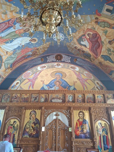 The image depicts the interior of a church, likely a Romanian Orthodox church, with extensive religious iconography. The ceiling is adorned with a mural depicting biblical scenes and figures, possibly angels and saints, rendered in a traditional style with vibrant colors and gold accents. A large, ornate brass chandelier hangs from the ceiling. The focal point of the lower section of the image is a wooden iconostasis, a wall of icons, separating the nave from the sanctuary. The iconostasis features intricately carved wooden details and numerous icons, including depictions of Jesus Christ, the Virgin Mary with the infant Jesus, and possibly saints or biblical events. Candles are visible in front of some of the icons, indicating active worship. A man wearing a light-colored shirt is partially visible in the foreground, looking downwards, suggesting he is participating in a service or observing the icons. Another person, partially visible from behind, wears a pink headscarf. The overall impression is one of a richly decorated and spiritually significant space. This specific imagery is consistent with the interior of the Saint Nicholas Church in Condrița, Moldova, known for its traditional Orthodox architecture and art.
