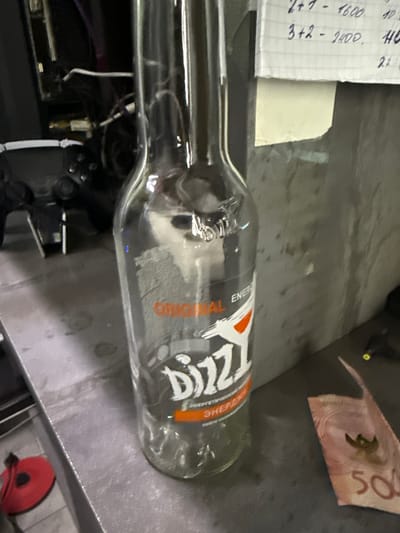 A clear glass bottle with an orange and white label is positioned vertically in the center of the frame, resting on a grey, metallic surface. The label on the bottle is partially obscured but appears to feature the word "DIZZI" in large, stylized white letters with an orange accent. The bottle is empty. To the left of the bottle, out of focus, are dark-colored electronic components and wires. To the right of the bottle, a crumpled piece of paper with handwritten notes is visible near the top edge of the frame. Below and to the right of the bottle, a fragment of a pink banknote with the number "500" visible is present on the grey surface. The background appears to be a textured, grey wall. The scene is indoors, with artificial lighting. No individuals are present in the frame.