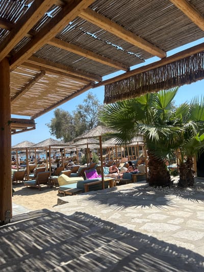 The image shows a beach scene in Piso Livadi, Greece. In the foreground, a wooden pergola with a thatched roof casts shadows on a stone patio. Beyond the patio, a sandy beach is lined with numerous lounge chairs and thatched umbrellas, many of which are occupied by people relaxing. Palm trees are visible on the right side of the image. In the background, the blue sea and sky are visible, along with some vegetation and other beach structures. The scene depicts a typical sunny day at a resort beach, with people enjoying leisure time under the shade of umbrellas and pergolas.