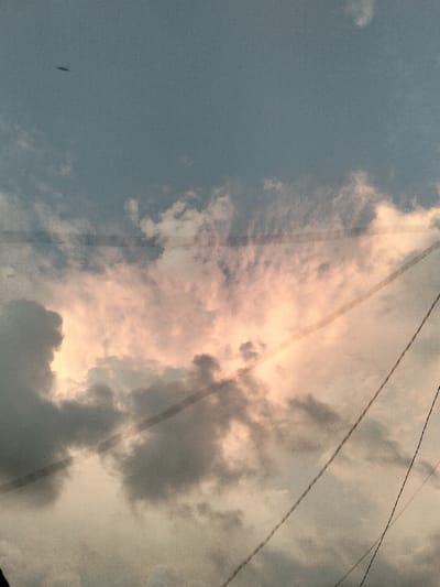 This image presents an overhead sky view, captured in the city of Panton, Indonesia. The sky is largely covered by clouds, displaying a gradient from muted blue-grey in the upper portion to warmer pink and orange hues in the lower-middle section, suggesting either dawn or dusk. Darker, dense grey cloud formations are prominent in the lower-left and central areas. Multiple dark, thin utility lines traverse the frame diagonally and horizontally, indicating an urban or suburban vantage point looking upwards. In the upper-left quadrant, a small, dark, elongated object is visible against the lighter blue sky, consistent with an unidentified aerial subject. No individuals, explicit actions, or direct interactions are discernible within the frame.