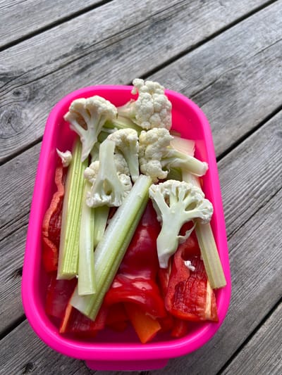 In Lommedalen, Norway, a bright pink plastic container is filled with a colorful assortment of chopped vegetables, including red bell peppers, celery sticks, and cauliflower florets. The container rests on a rustic wooden surface composed of weathered gray planks with visible grain and occasional knots. The overhead perspective offers a clear view of the ingredients, suggesting they are prepared for a snack or meal. The lighting is natural and diffused, casting subtle shadows and highlighting the fresh textures of the vegetables.