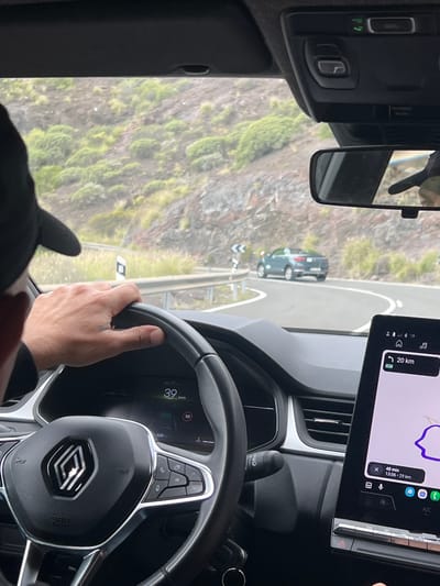 The image, taken from the driver's seat, captures a scene of driving along a winding road in Mogán, Spain. The driver's hands are on the steering wheel of a Renault, indicated by the logo. The dashboard screen shows a speed of 39 km/h. A navigation app is displayed on a tablet mounted on the dashboard, indicating the destination is 20 km away and the estimated time of arrival is 48 minutes later at 13:06. Outside the window, the landscape reveals a mountainside covered with greenery. A vehicle is visible on the road ahead.