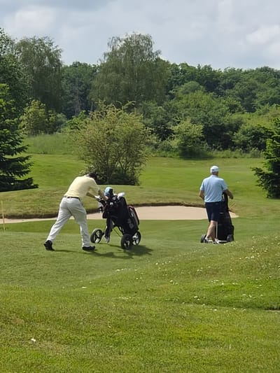 Two men are playing golf on a green course in Lenningen, Luxembourg. One man in a yellow shirt and white pants is reaching into a golf bag on a black golf cart. The other man is wearing a light blue shirt and dark blue shorts, pulling his own golf bag on a cart behind him. Trees surround the golf course.