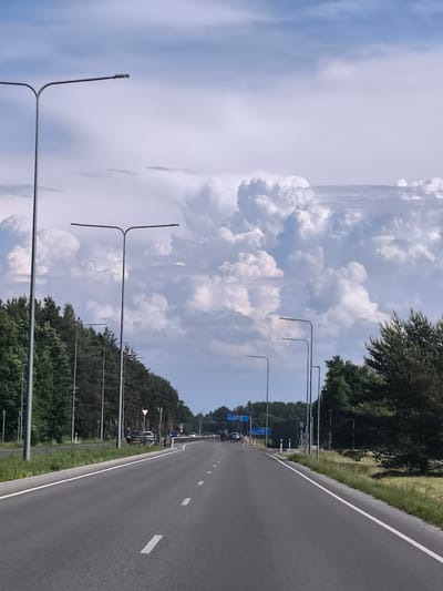 A straight, empty asphalt road stretches into the distance under a sky filled with dramatic, white cumulus clouds against a pale blue backdrop. The road is lined on both sides with tall, slender streetlights, their sleek, modern design contrasting with the natural scenery. On the left, a dense line of tall, green pine trees forms a natural barrier, with some grass and smaller bushes visible at their base. On the right, the trees are also dense, with a small clearing of dry grass and some darker, bushier vegetation closer to the road. The road is marked with white dashed lines, indicating traffic lanes. In the distance, a few vehicles are visible, some on the road and others parked on the shoulder. The overall scene suggests a rural or semi-rural highway environment in Tiskre, Estonia, on a partly cloudy day.