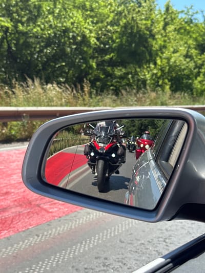 The image is a rearview mirror reflection of a motorcycle convoy on a road, likely in or near Prisovo, Bulgaria. A silver car's side mirror is visible in the foreground, reflecting a road with a bright red stripe on the left side. In the reflection, a black and red motorcycle with saddlebags is the closest object, followed by other motorcycles behind it, one of which is red. The motorcycles appear to be traveling in the same direction as the car. The background outside the mirror shows lush green trees and foliage, suggesting a rural or semi-rural setting. The sky is bright blue, indicating a clear, sunny day. The road surface has a patterned texture with lighter spots, possibly from sunlight reflecting off it.
