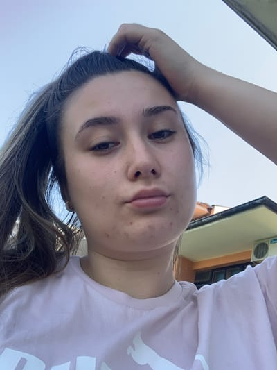 A low-angle, close-up selfie shows a young woman with her hand on the back of her head, looking at the camera with pursed lips. She has brown hair pulled back in a ponytail and is wearing a light pink t-shirt. The background appears to be an outdoor setting with a clear blue sky and the side of a building with an air conditioning unit visible. The image was captured in Chernomorets, Bulgaria.