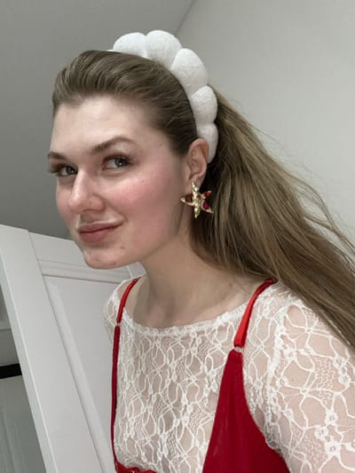 A woman faces the camera, smiling slightly. She has light brown hair and wears a white, puffy headband. A gold, jeweled star earring adorns her ear. She is dressed in a white lace top with red straps and accents. The background consists of a white wall and the corner of a white cabinet or door. The image is slightly rotated.