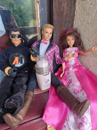Three fashion dolls and a perfume bottle are arranged vertically on a multi-textured surface, likely outdoors on a bright, sunny day.
At the top, a female doll with long brown hair (featuring pink streaks) and blue eyes wears an elaborate pink and white princess-style gown with silver floral patterns.
Below her is a male doll with blonde hair and blue eyes, dressed in a black and pink plaid jacket over a white shirt with a pink bow tie. His arm is positioned around a light pink cylindrical perfume bottle. The bottle's label clearly reads "HAYA" and "LE CHAMEAU EAU DE PARFUM" in English, accompanied by Arabic script, and its cap is adorned with an ornate pattern.
At the bottom, another male doll wears dark hair and green-tinted sunglasses. He is clad in a black hooded outfit featuring a circular chest logo with an orange, stylized "M" or "W" on a black and grey background. He also wears black gloves with yellow cuffs and brown boots.
The dolls and bottle rest upon various surfaces: rough grey concrete at the upper left, a dark wooden plank in the middle, and a reflective surface (possibly glass) at the bottom left showing distorted reflections of green foliage and sky. A reddish-brown, textured surface, resembling leather or fabric, is visible on the right. The arrangement appears staged in what seems to be an outdoor or semi-outdoor setting.