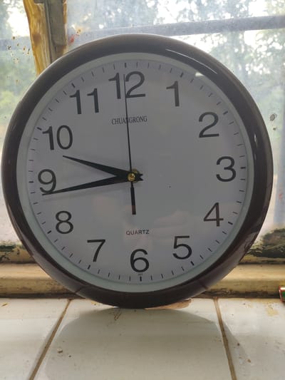 A dark-rimmed, circular analog clock with a white face is displayed. The clock has black hour and minute hands, with the hour hand pointing slightly past the 9 and the minute hand pointing to the 10. The numbers 1 through 12 are clearly visible in black. The clock is positioned on a ledge, possibly a windowsill, against a backdrop of a hazy, out-of-focus window with sunlight filtering through. The environment appears somewhat weathered, with visible grime and dust on the ledge and the window frame. The background suggests an outdoor setting with foliage. The clock face reads "CHUANGRONG" and "QUARTZ".