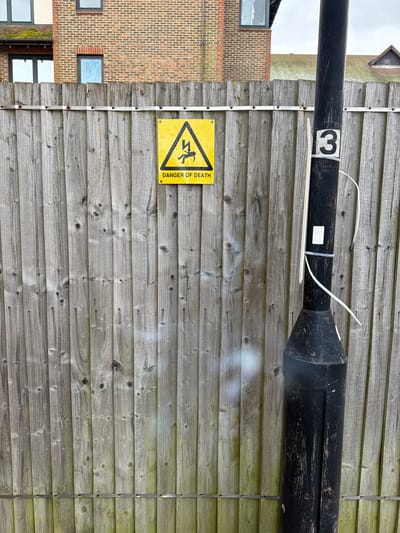 The image shows a wooden fence in Mole Valley, United Kingdom. Affixed to the fence is a yellow triangular "Danger of Death" sign depicting a person being electrocuted. A black, cylindrical object with a number "3" on it, possibly a repurposed piece of industrial equipment, is mounted horizontally along the upper portion of the fence. A building constructed of red and orange brick is partially visible to the left of the fence. The weather appears overcast.